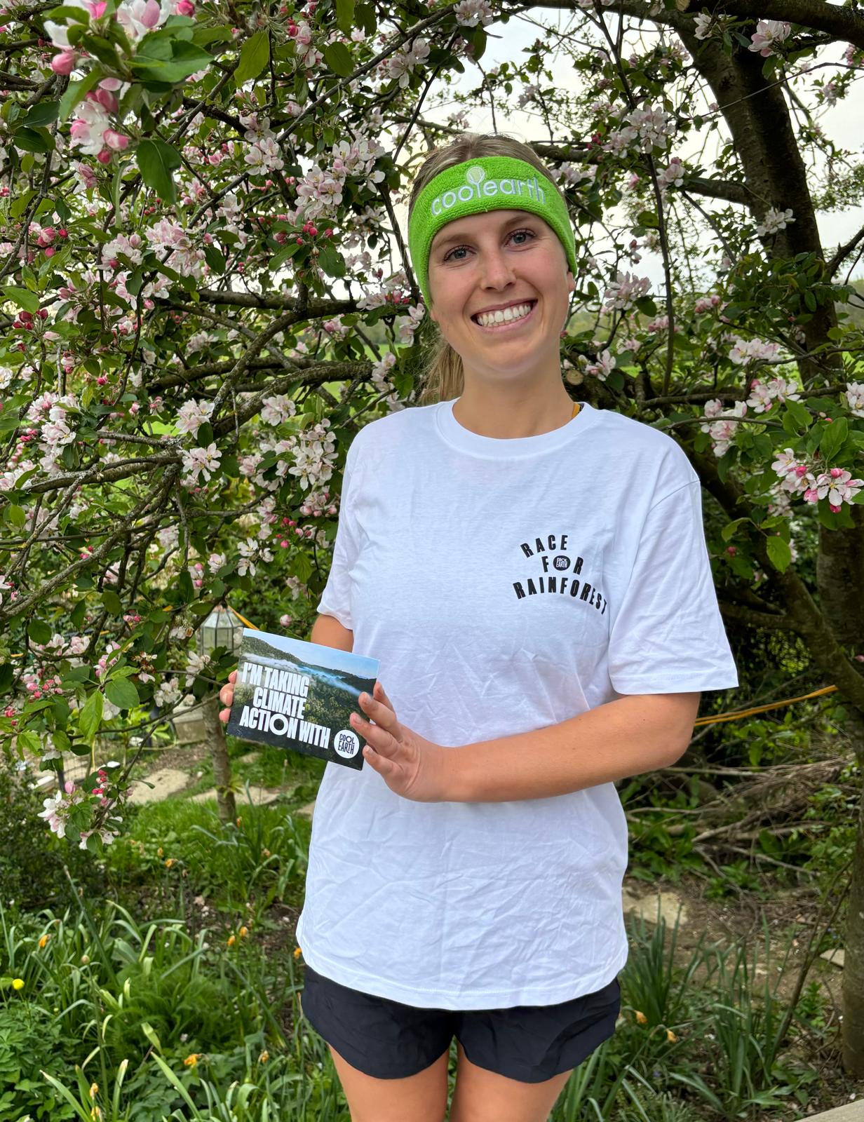 Fundraiser Ebony stands in front of blossom covered tree. She sports a white Cool Earth tshirt and green headband and a big smile.